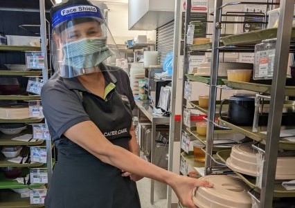 Cater Care aged care team member in a mask and face shield taking a tray from the back area.