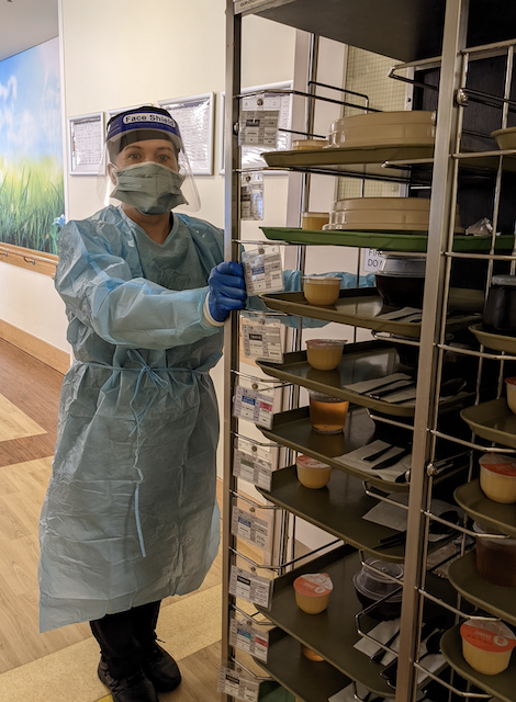 Cater Care aged care team member is in full protective gear (mask, face shield and gown), while getting ready to take meals to residents via the tray trolley.