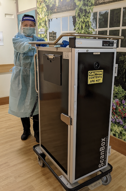 Cater Care aged care team member is in full protective gear (mask, face shield and gown), while getting ready to take meals to residents via the heated trolley.